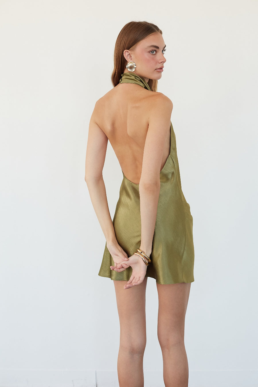 A woman modeling a halter-neck mini dress with an open back, shown from the back. The dress has a shiny olive green satin-like finish and is photographed against a plain white studio background.