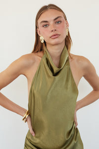 A woman modeling a sleeveless olive green halter-neck top with a draped cowl neckline, paired with gold earrings and a gold bangle, against a plain light background.