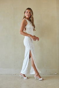 Woman wearing a white lace dress with a slit standing against a plain background