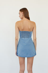 A woman shown from the back wearing a strapless light blue denim mini dress with a smocked bodice, set against a plain white studio background.