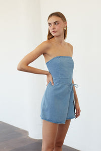 A woman modeling a strapless light blue denim mini dress with a tie detail at the waist, standing against a plain white wall.
