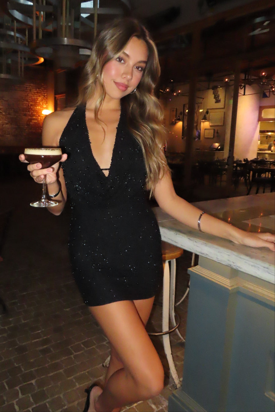 girl in a black embellished cowlneck mini dress standing at the bar with a drink