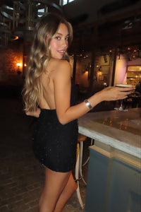 girl in a black embellished mini dress standing at the bar with a drink showing off the back of the dress