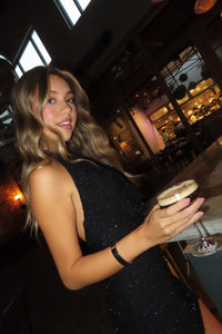 girl in a black embellished mini dress standing at the bar with a drink
