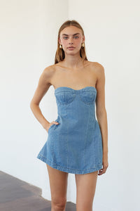 A woman modeling a strapless light blue denim mini dress with a sweetheart neckline and a flared skirt, standing against a plain white wall.