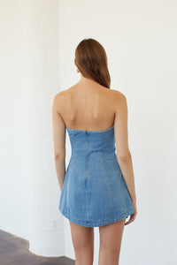 A woman viewed from behind wearing a strapless light blue denim mini dress with a structured corset-style bodice and flared skirt, standing against a plain white wall.