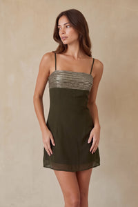 A woman modeling a short olive green spaghetti-strap dress with a metallic silver sequined bodice, set against a neutral beige textured studio backdrop.