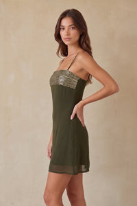 A woman modeling a short olive green spaghetti-strap dress with a metallic silver sequined bodice, set against a neutral beige textured studio backdrop. Side angle