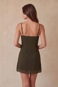 A woman modeling a short olive green spaghetti-strap dress, set against a neutral beige textured studio backdrop.