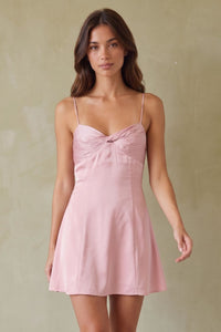 pink-image | A woman wearing a light pink mini dress with thin spaghetti straps and twisted bodice, photographed against a textured beige wall.