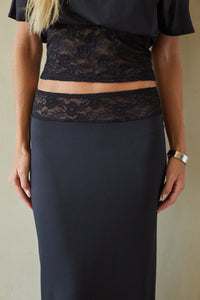 A model wearing a black midi skirt with a sheer floral lace waistband and a matching black top, shown from the chest down against a neutral beige background.