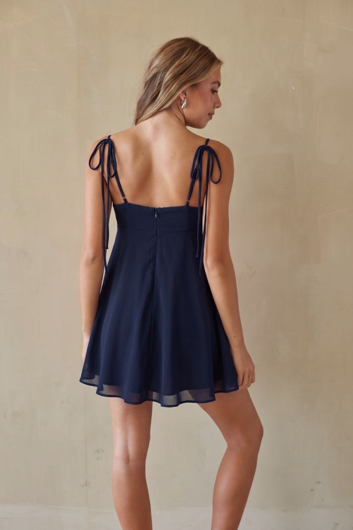 A woman modeling a navy blue sleeveless mini dress from the back, featuring thin tie straps and a flared skirt, set against a neutral beige textured wall.