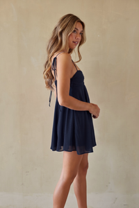 A woman modeling a short navy blue mini dress with thin shoulder ties, standing against a textured beige wall.