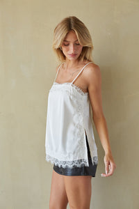 A woman wearing a white lace-trim camisole top and dark denim shorts, standing against a textured beige wall.