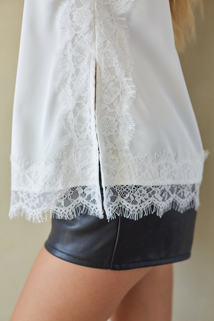 Close-up of a white blouse with intricate lace trim and a split back, worn by a model with black faux-leather shorts.