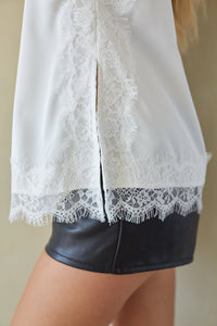 Close-up of a white blouse with intricate lace trim and a split back, worn by a model with black faux-leather shorts.