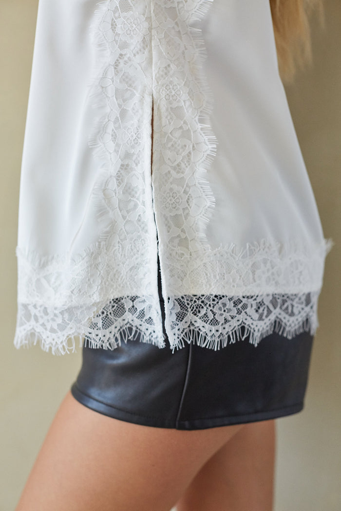 Close-up of a white blouse with intricate lace trim and a split back, worn by a model with black faux-leather shorts.