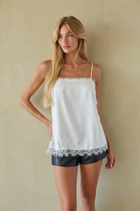 A blonde model wearing a white spaghetti-strap camisole top with delicate lace trim and black faux-leather shorts, standing against a textured beige wall.