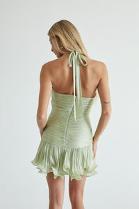 A woman shown from the back wearing a light sage green halter mini dress with a ruffled hem, photographed against a plain light gray studio background.