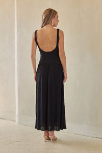 A woman modeling a black sleeveless maxi dress with a square neckline and a flowing skirt, standing against a neutral beige wall.