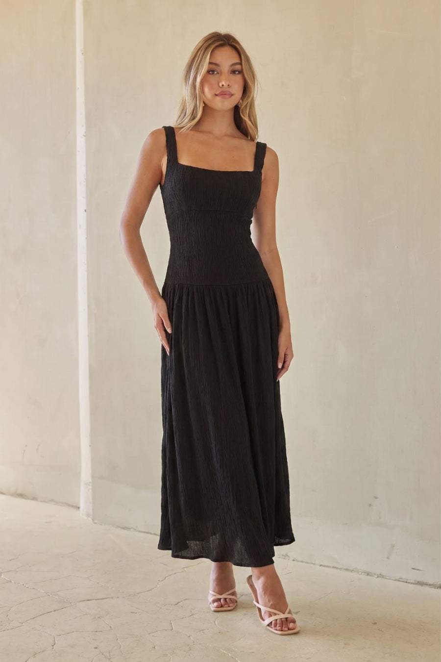 black-image | A woman modeling a black sleeveless maxi dress with a square neckline and a flowing skirt, standing against a neutral beige wall.