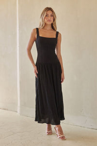 black-image | A woman modeling a black sleeveless maxi dress with a square neckline and a flowing skirt, standing against a neutral beige wall.