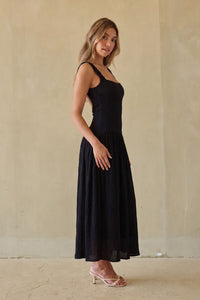A woman modeling a black sleeveless maxi dress with a fitted bodice and flowing skirt, standing against a textured beige wall.