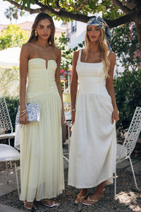 Two women in white dresses standing outdoors with trees and chairs in the background.
