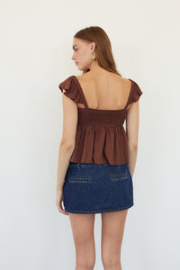 A woman shown from the back wearing a brown ruffled off-the-shoulder peplum top and a blue denim mini skirt, standing against a plain white background.