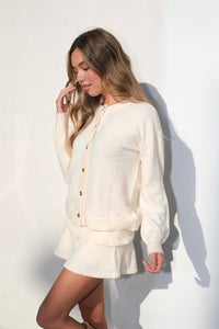 girl against a white wall wearing a cream colored cardigan and mini skort set.