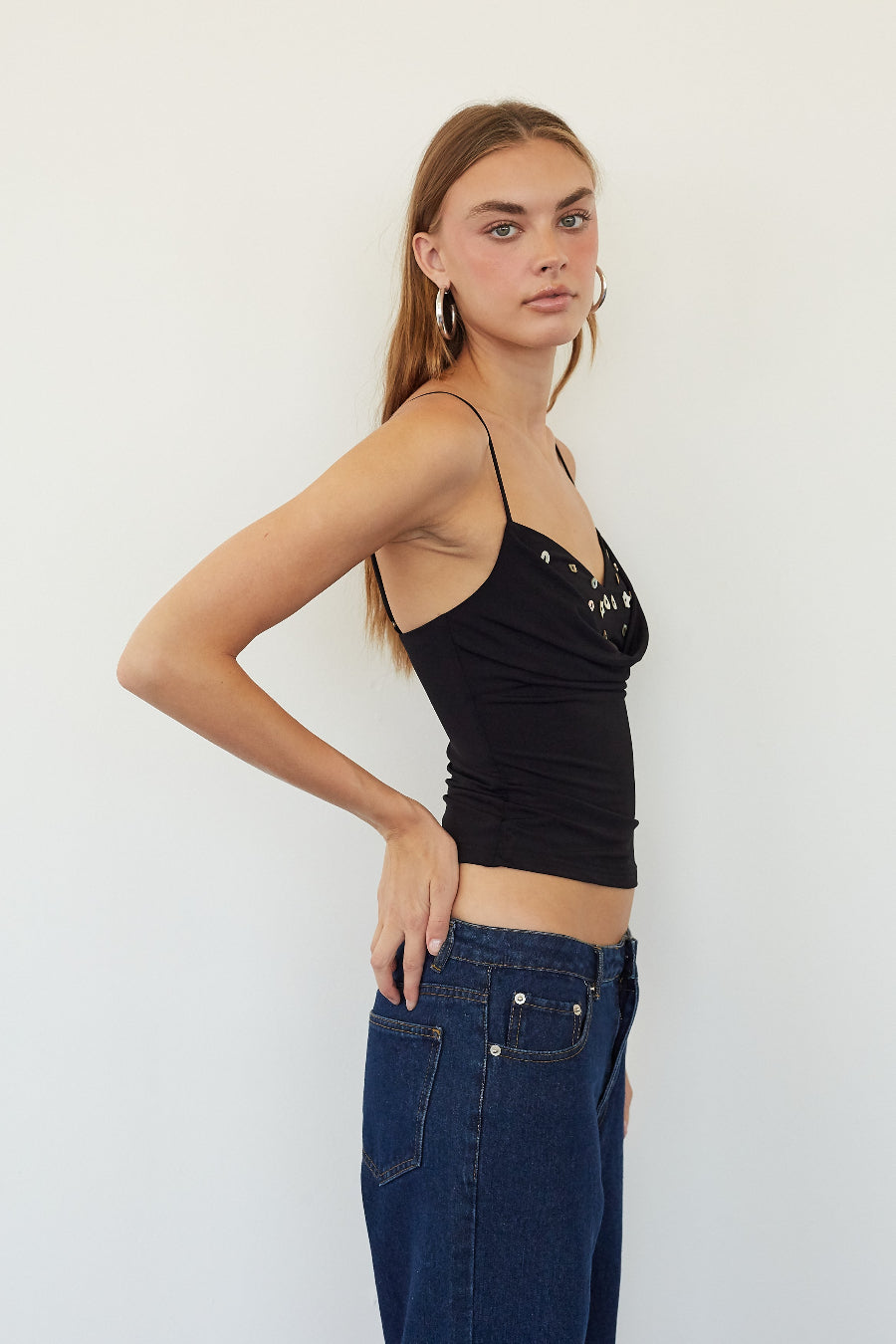 A woman modeling a black spaghetti-strap crop top with silver rhinestone embellishments on the cups, paired with dark blue jeans, against a plain white studio background.