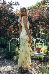 Woman in a floral dress standing outdoors with trees and a chair in the background
