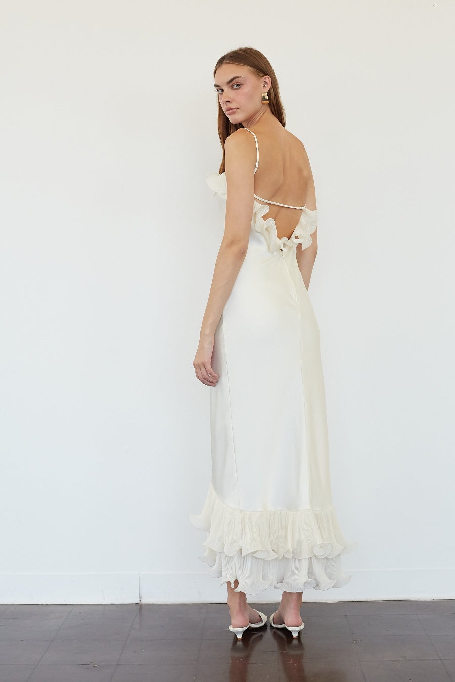 A woman modeling a long cream maxi dress from the back, featuring thin straps, a ruffled open back, and a tiered ruffle hem, set against a plain white wall.