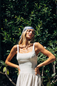 Woman in a white dress with a blue headscarf standing against a green leafy background