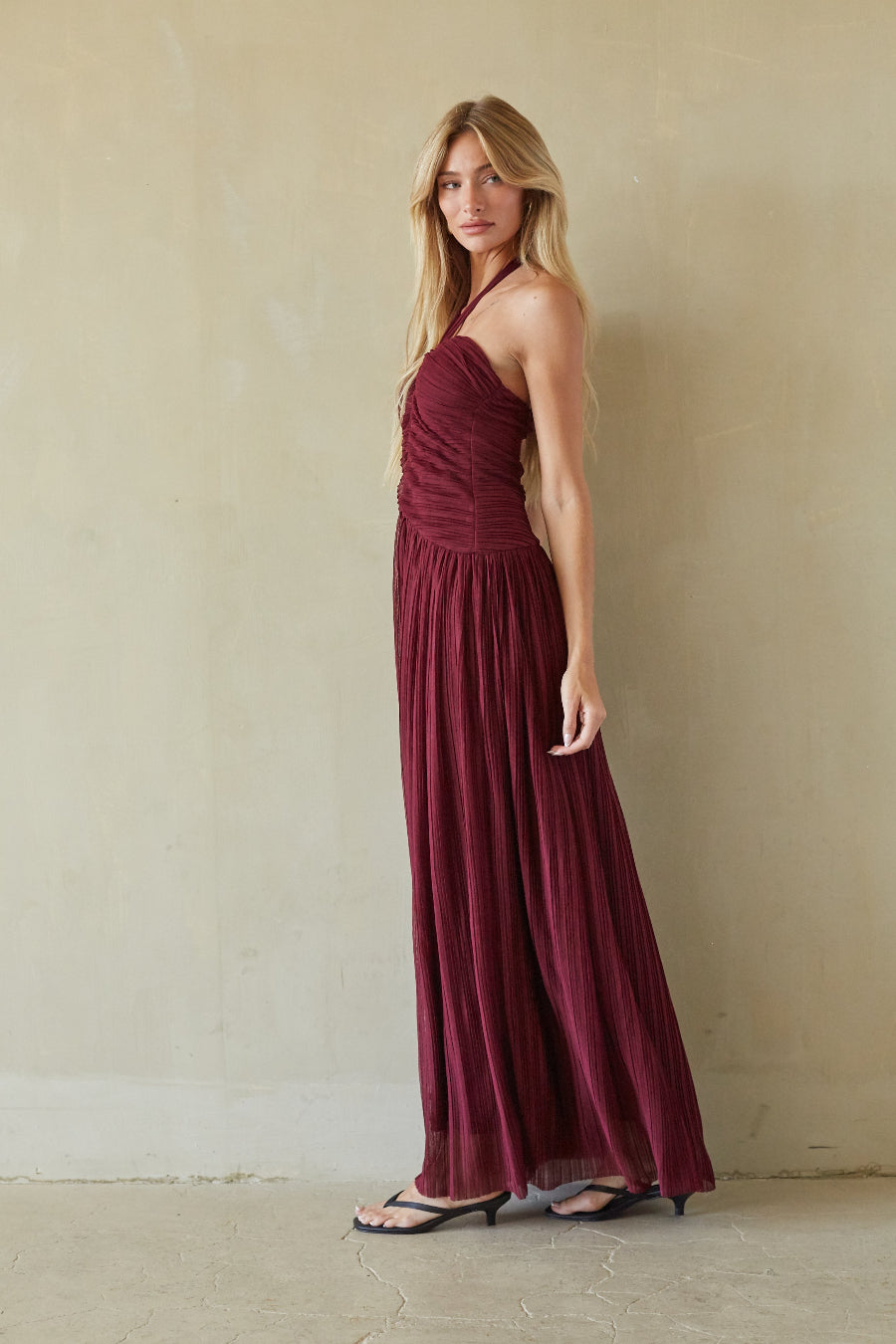 A woman modeling a floor-length burgundy halter maxi dress with a ruched bodice and flowing pleated skirt, standing against a textured beige wall.