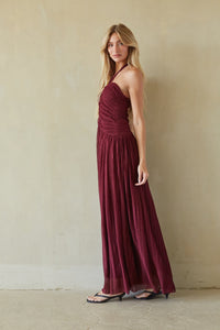A woman modeling a floor-length burgundy halter maxi dress with a ruched bodice and flowing pleated skirt, standing against a textured beige wall.