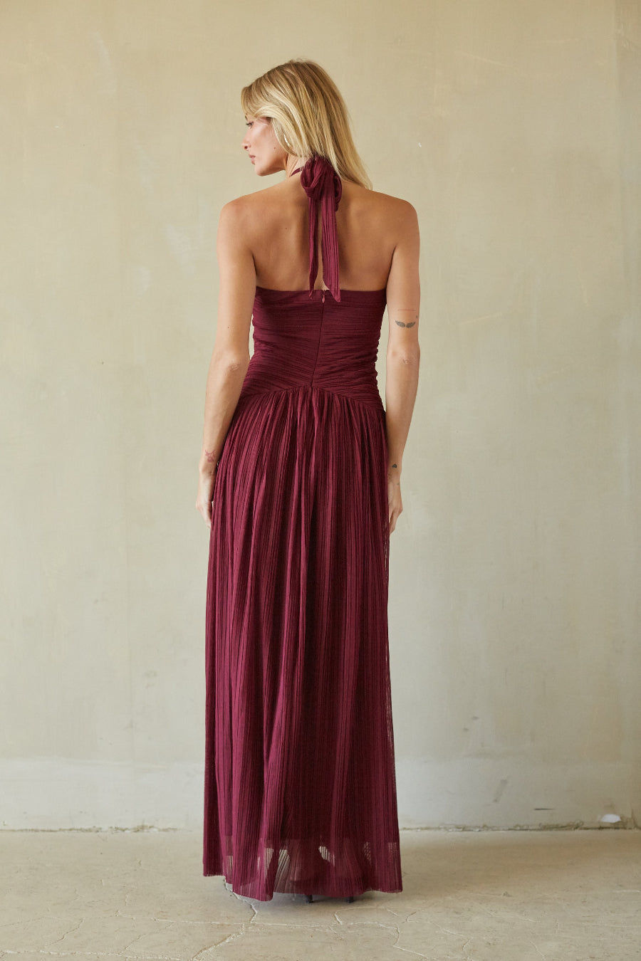 A woman wearing a sleeveless, back-tie maxi dress in deep burgundy, photographed from behind against a neutral beige plaster wall.