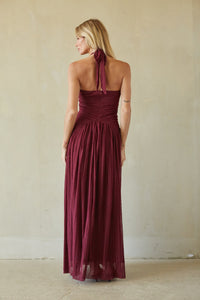 A woman wearing a sleeveless, back-tie maxi dress in deep burgundy, photographed from behind against a neutral beige plaster wall.