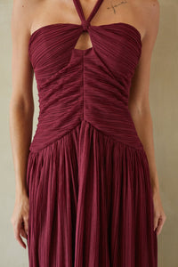 A woman wearing a sleeveless halter-neck maxi dress in deep burgundy with a pleated texture, featuring a ruched bodice and a keyhole cutout at the chest.