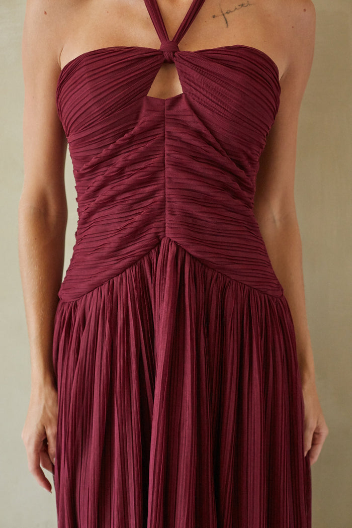A woman wearing a sleeveless halter-neck maxi dress in deep burgundy with a pleated texture, featuring a ruched bodice and a keyhole cutout at the chest.