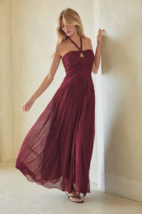 A woman modeling a sleeveless, halter-neck maxi dress in deep burgundy. The dress features a ruched bodice and a flowing, pleated skirt, shown in a soft, warm-toned indoor setting with beige walls.