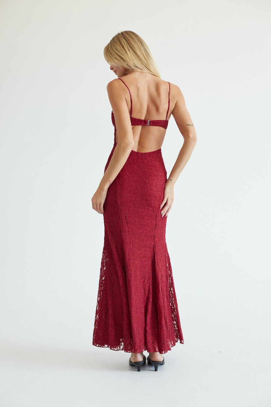 A woman viewed from the back wearing a long burgundy lace maxi dress with thin straps and an open back, standing against a plain light gray studio background.