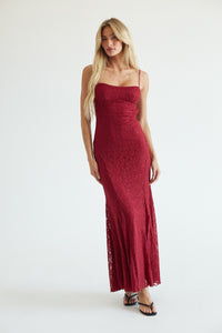 red-image | A blonde model wearing a sleeveless, floor-length burgundy lace maxi dress with thin straps and a fitted silhouette, standing against a plain white studio background.