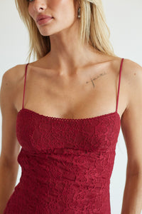 A close-up of a woman wearing a burgundy lace camisole top with thin spaghetti straps and a scoop neckline, set against a plain light background.