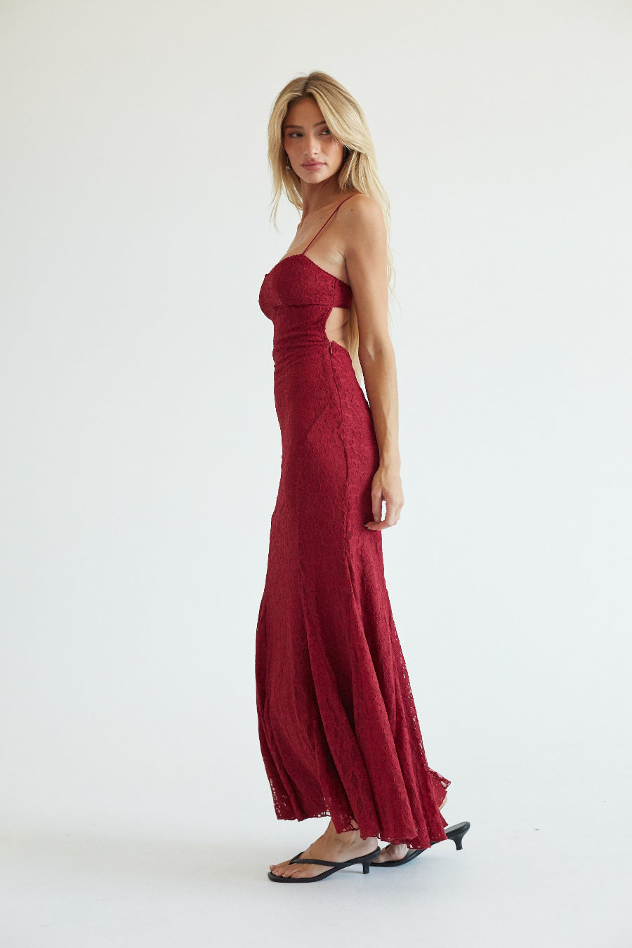 A blonde model wearing a sleeveless, floor-length burgundy lace maxi dress with thin straps and a fitted silhouette, standing against a plain white studio background.