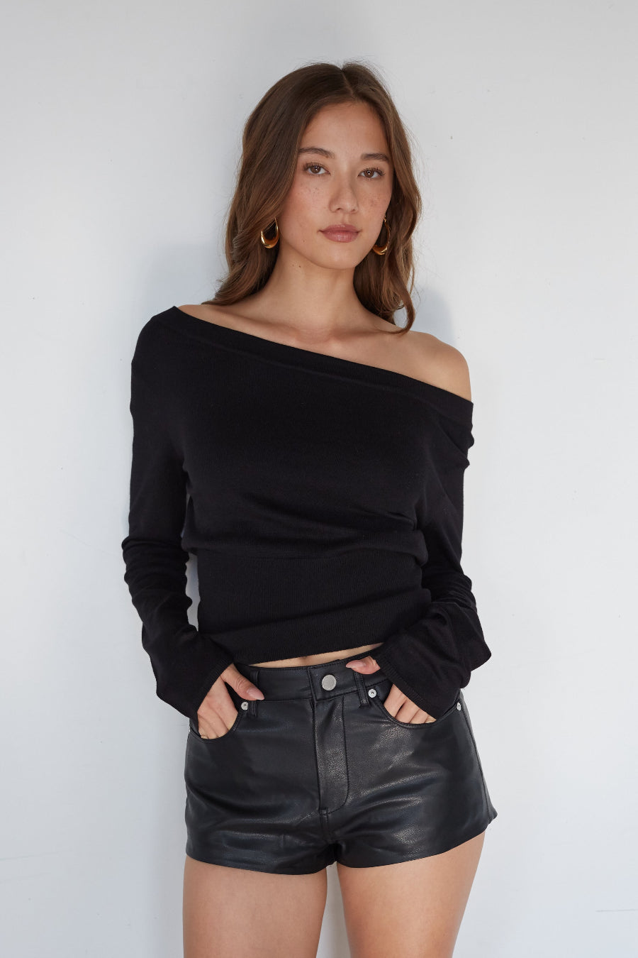 Woman wearing a black off-shoulder top and black leather shorts against a white background