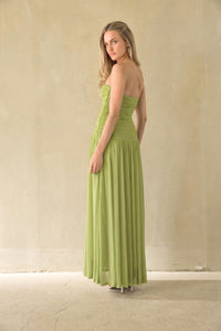 A woman modeling a strapless lime green maxi dress with a ruched bodice and flowing skirt, standing against a textured beige wall.