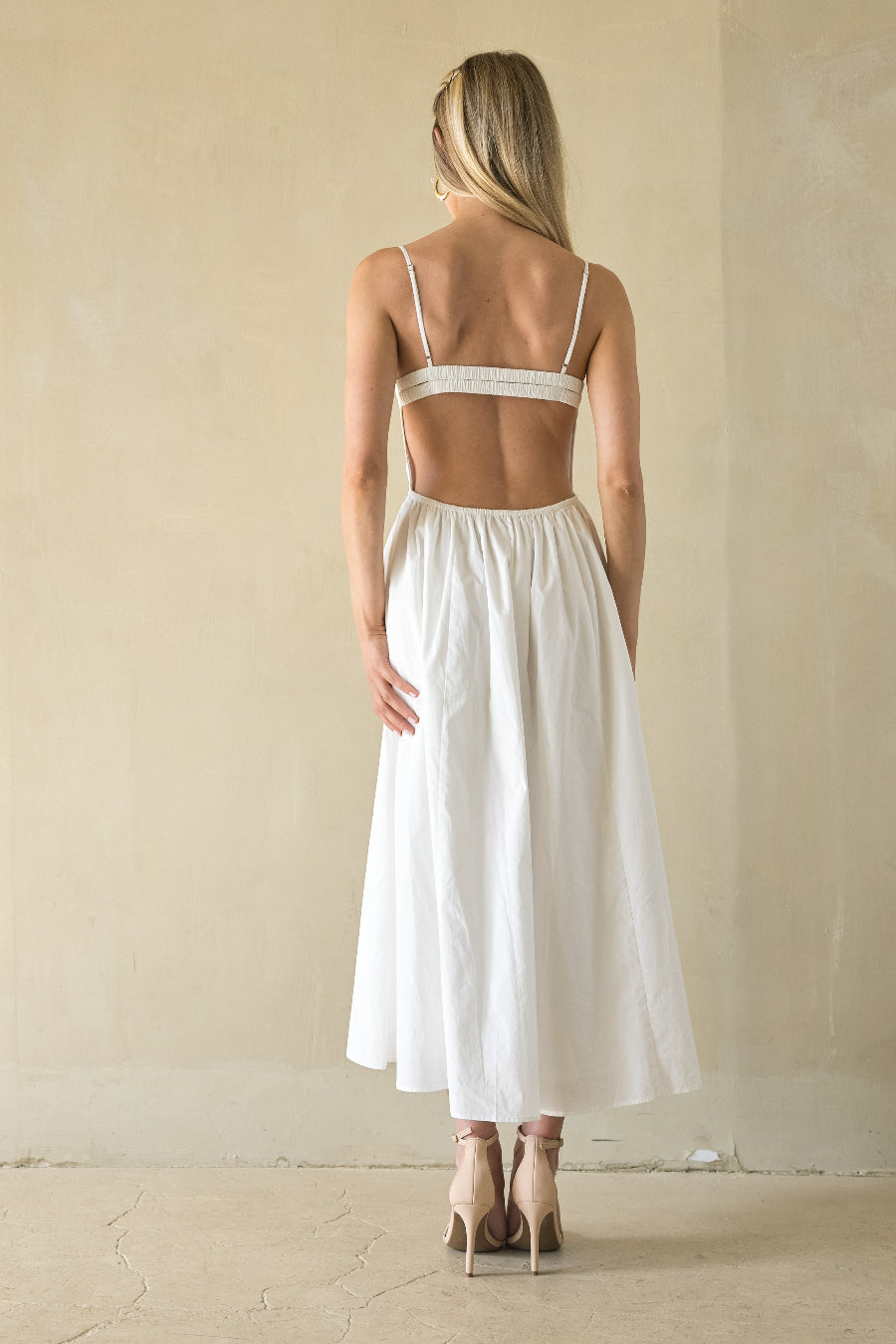 Woman wearing a white dress with thin straps and open back in a minimalistic setting.