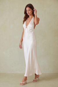 A woman modeling a white satin halter maxi dress with a side slit, paired with white wedge sandals, standing against a plain beige wall.