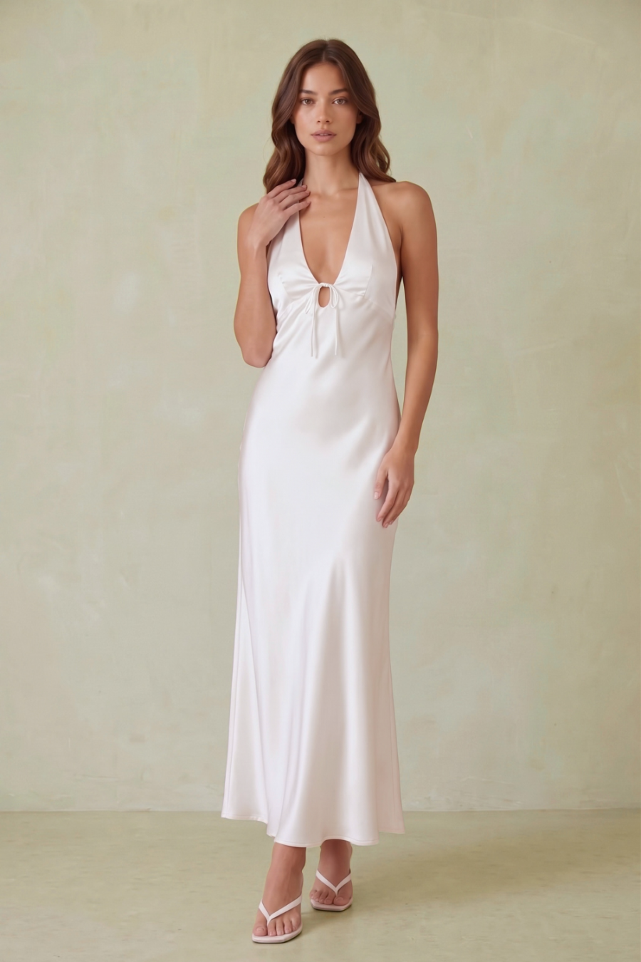 A woman modeling a white satin halter maxi dress with a keyhole cutout at the bust, standing against a textured beige studio backdrop.